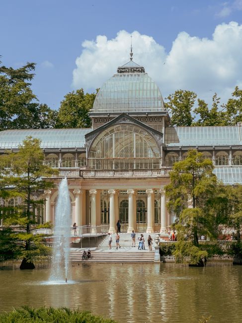 A large historic glass and iron greenhouse structure with intricate metal framing and multiple domed glass sections, situated outdoors with clear daylight. The building features a prominent central do