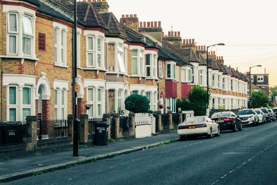 A street scene on Westow Street in Crystal Palace, showing a row of parked cars lining both sides of the road, with storefront shops and buildings, including a pharmacy and other businesses, on the ri